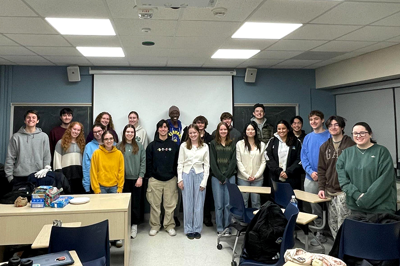 A group of 21 college students stand inside a classroom for a group photo.