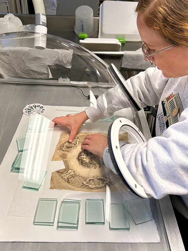 A student carefully flattens a 19th century transferware print that sits on a table inside a clear dome.