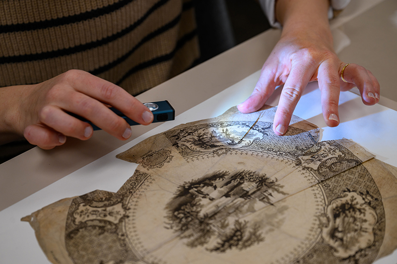 A close-up view of a student's hands holding a light to inspect a 19th century transferware print.