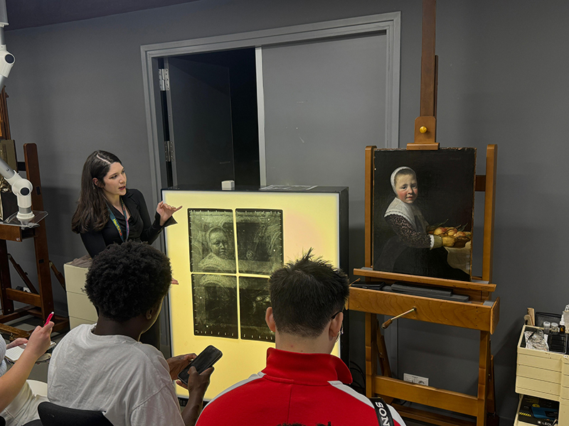 A graduate student in a classroom presents x-radiographs of an adjacent 17th century painting.