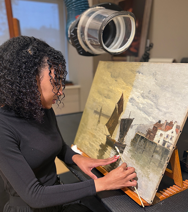 A graduate student in an art studio restores a 1965 painting on an easel.