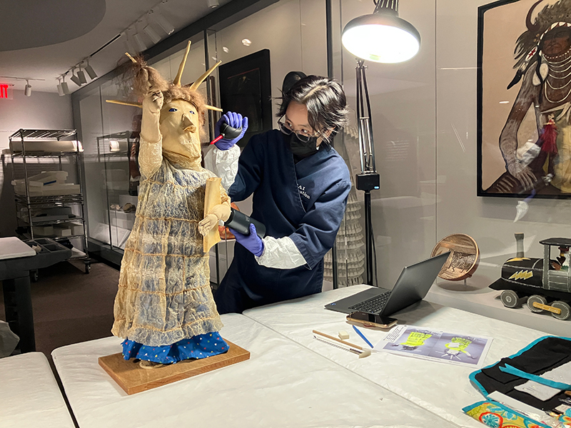 A graduate student wearing a face mask and blue latex gloves in an art laboratory treats a fabric sculpture.