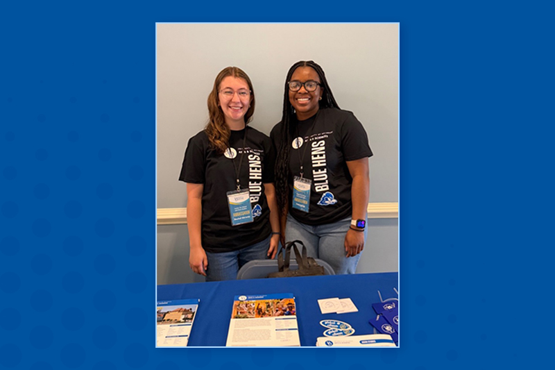 Two college students in black T-shirts stand in front of a display table with brochures and flyers.