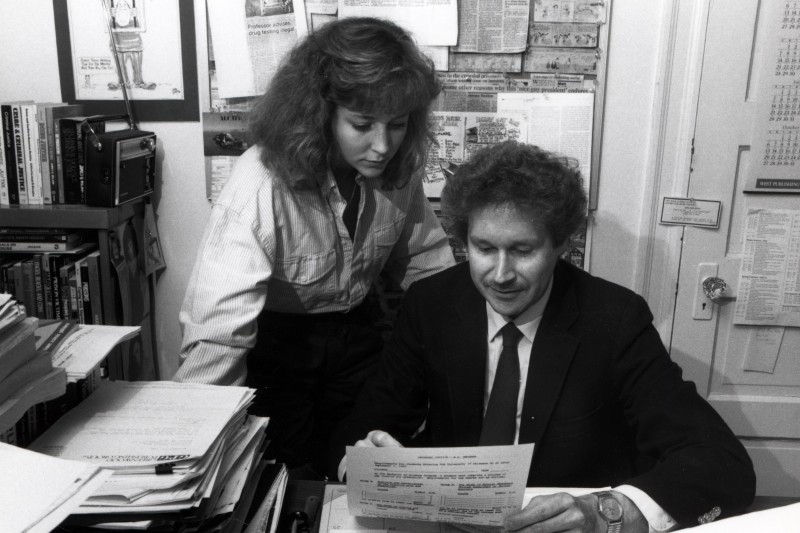 Black and white photo of a college professor in his office reading a document with a student standing behind him.
