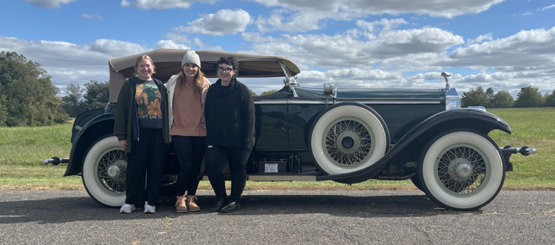 Three college students standing in an open field in front of a green 1927 Rolls Royce.