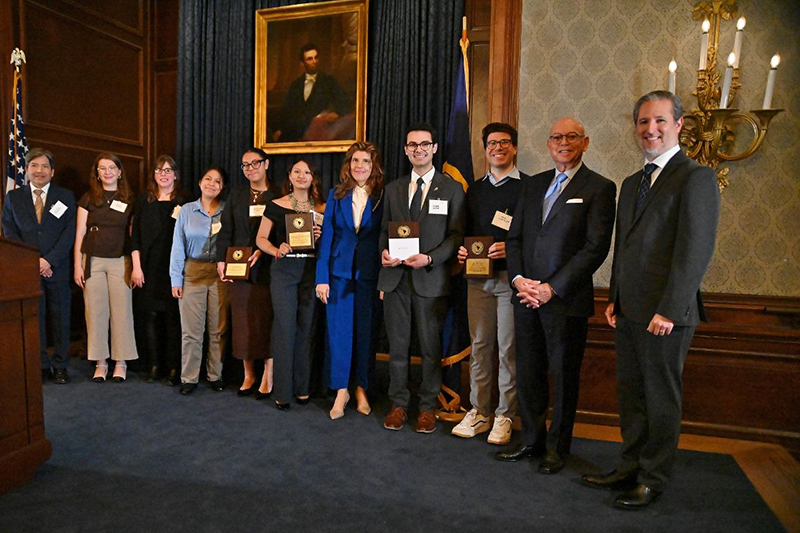 A group of students holding plaques and award representatives, dressed in formal business attire, inside room with period furnishings and an Abraham Lincoln portrait.