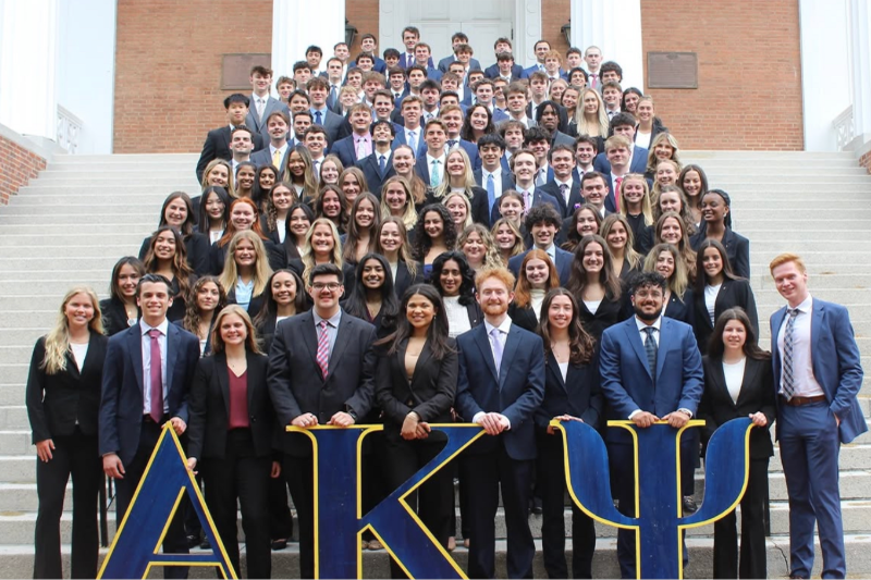 A large group of college students in business attire stand for a group photo on the steps of Memorial Hall behind large blue fraternity letters for Alpha Kappa Psi.