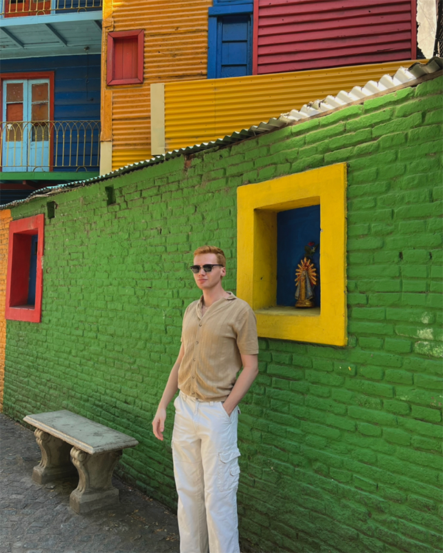 A college student stands next to a colorful green, yellow and orange brick home in Granada, Spain.