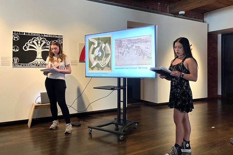 Two college students inside an art gallery deliver a presentation while standing next to a monitor with a slide presentation.