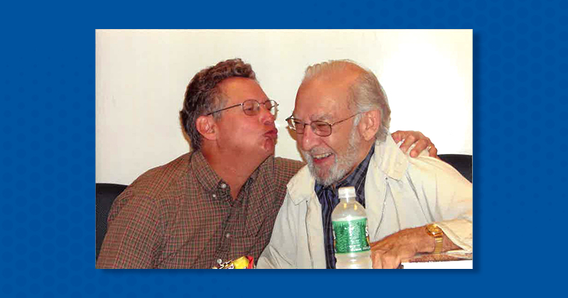 A college professor laughs as another professor embraces and kisses him.