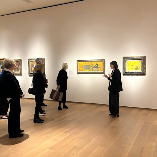 A woman in an art gallery stands in front of paintings and addresses museum patrons.