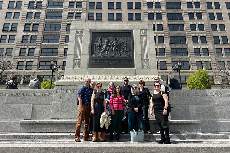 A group of 10 people stand in front of a large monument in a city on a sunny day.