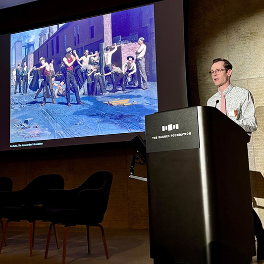 A college student delivers remarks from behind a podium while standing next to a slide presentation of a 19th century painting.