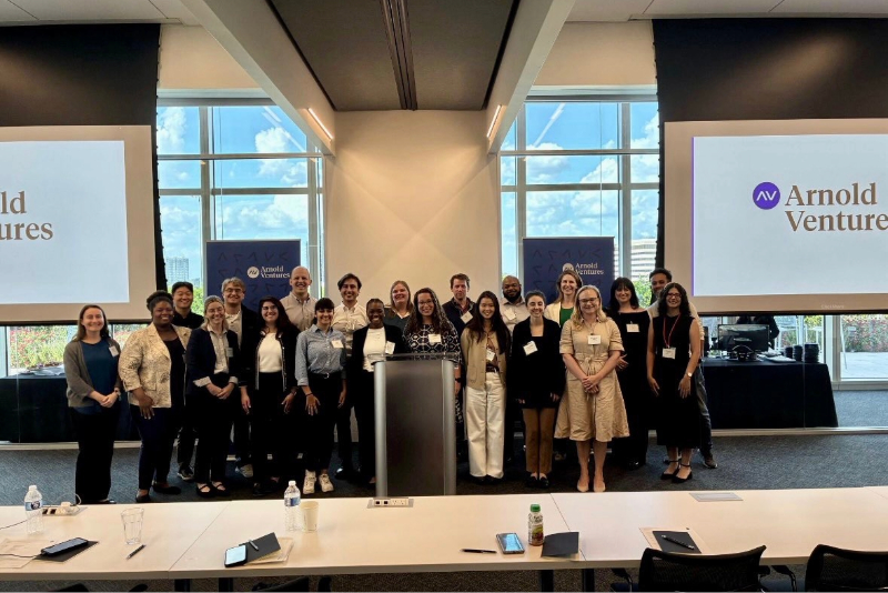 A group of smiling students gather for a group photo in the front of a conference room.