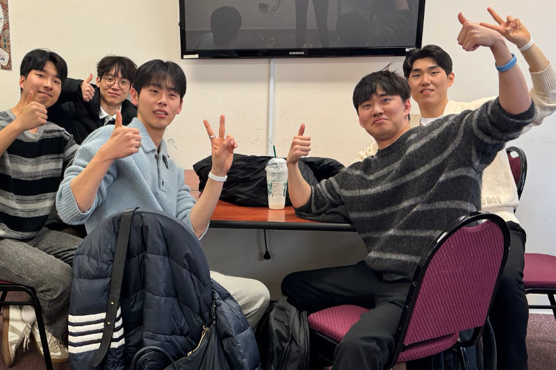 5 male students sit at a table in their Reading Cluster class and throw up peace signs.