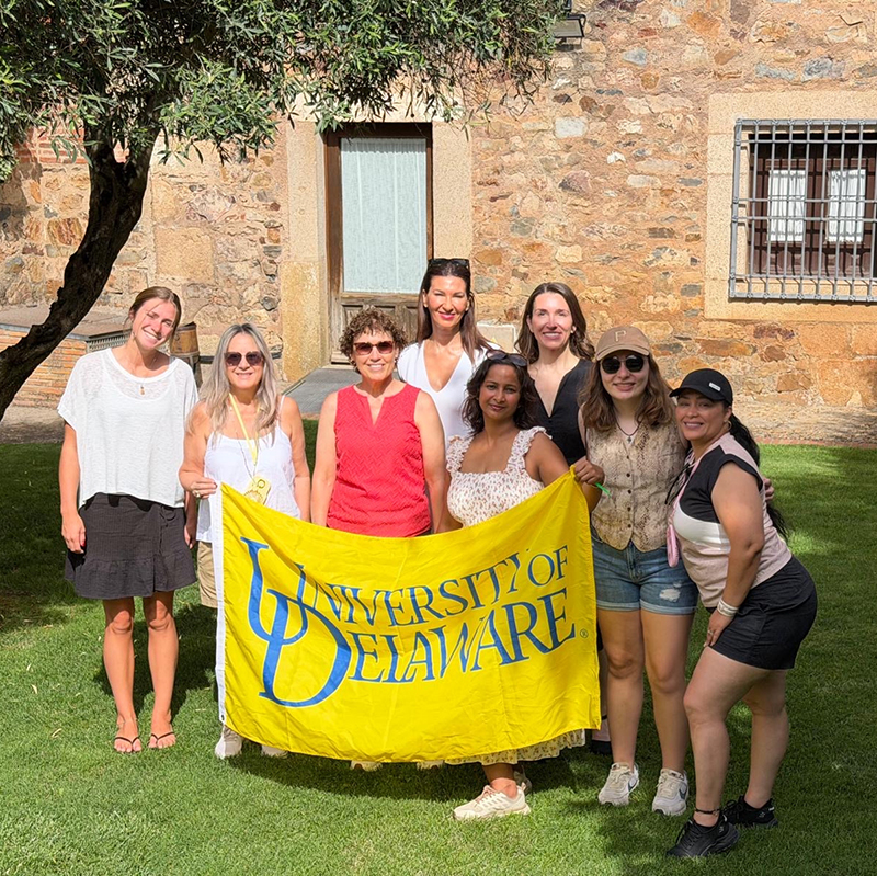 A group of people standing outside on a sunny day holding a University of Delaware banner.