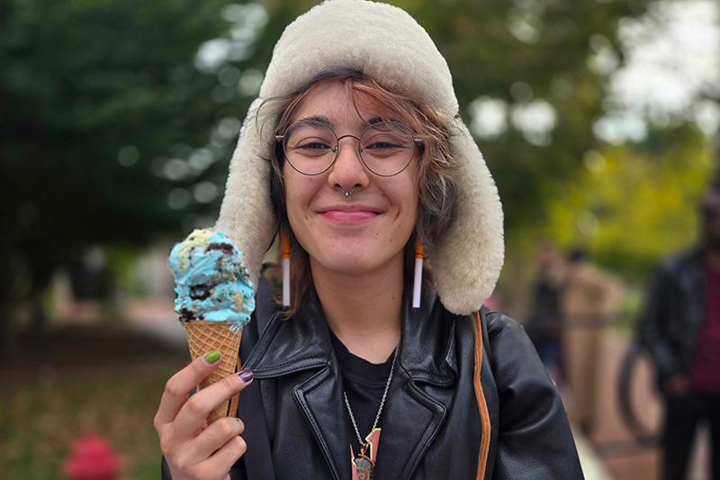 A college student in furry hat holds an ice cream cone outside on a college campus.