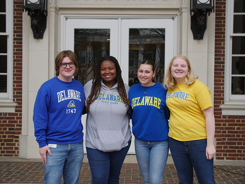 4 students wearing UD gear posing in front of campus building