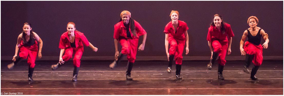 Six people dressed in red cultural wear dancing on stage performing South African gumboot dance