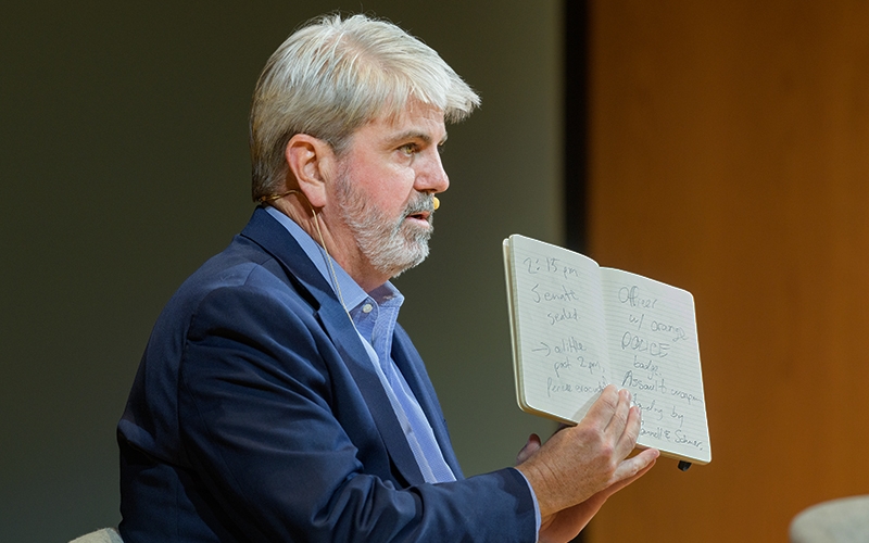Guest speaker on stage inside a theatre at the University of Delaware addresses the audience while holding up his notebook.
