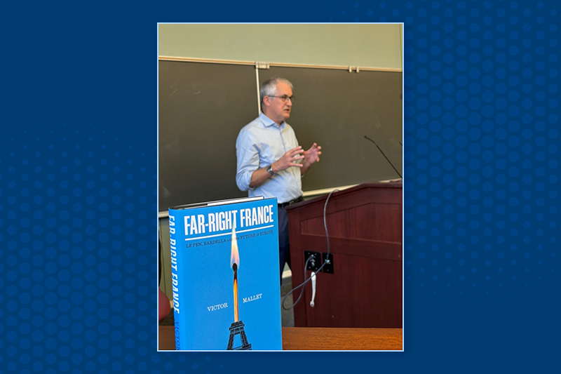 An author in a classroom delivers remarks from a podium with a book cover of 