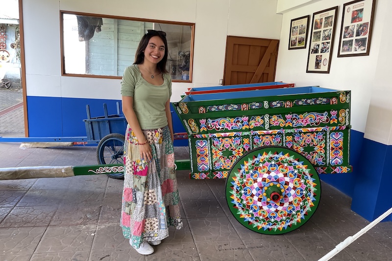 College student Amber Tiongson, dressed in a festive long skirt, stands next to a brightly painted cart.