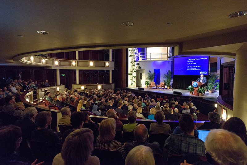 An audience listens inside a University of Delaware theater listens to a guest speaker who stands on stage behind a podium and delivers a presentation with Powerpoint slides.