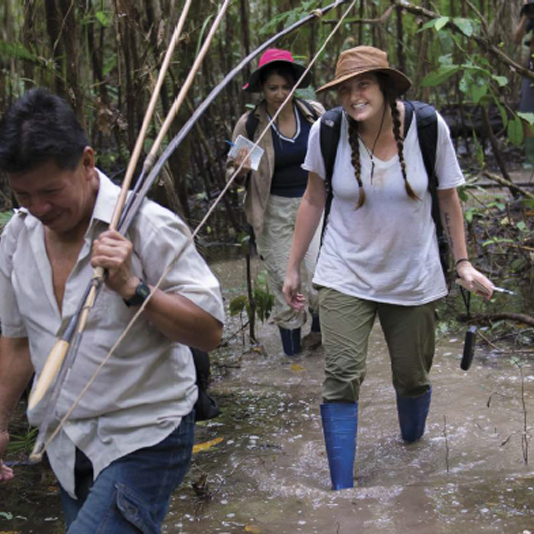 Chelsea Rozanski wears a safari hat and blue rubber boots and walks through a muddy stream in a tropical forest, following a man carrying a bow and arrows.