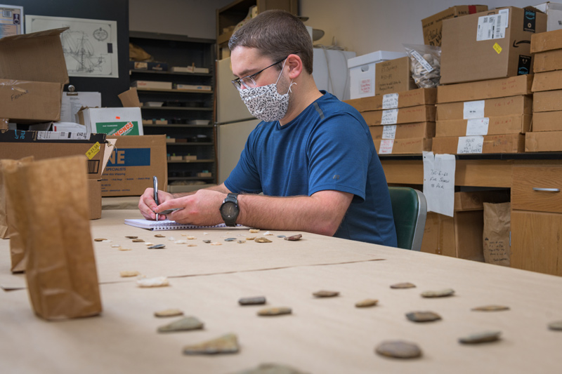 University of Delaware student Nicholas Gonnella sits inside a research laboratory at a table covvers with arrowheads, spear points and ceramics and writes notes in a notebook.