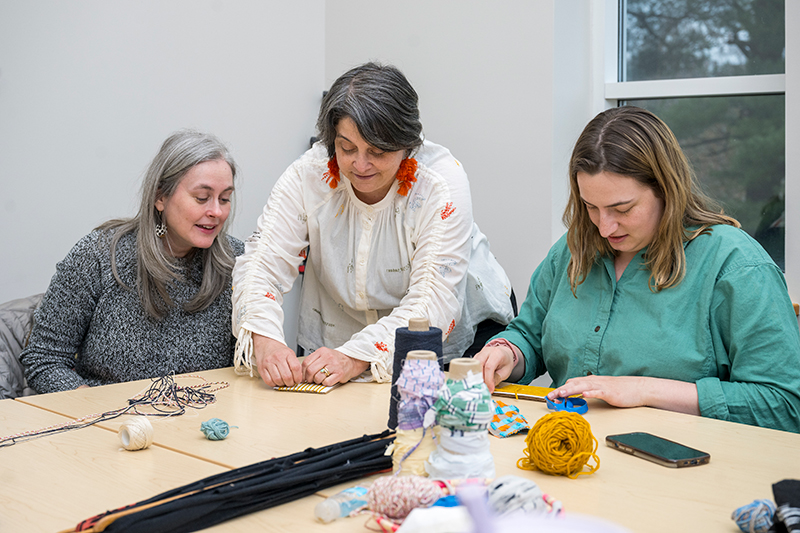 Two University of Delaware professors and a graduate student sit at a table with colorful textiles and yarn  and work together on rag projects.