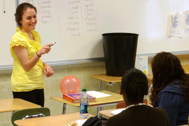 A student teacher stands in front of a classroom, writes notes on a whiteboard, and leads a class discussion with students seated at their desks. 