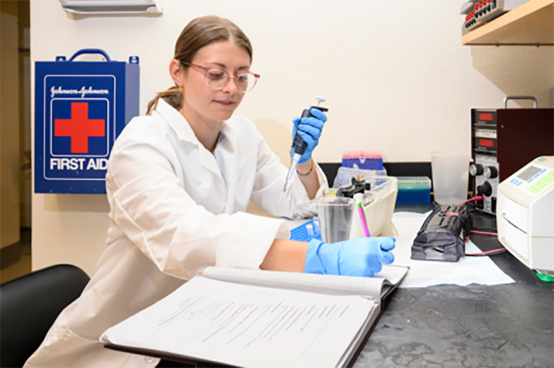 College student wearing blue rubber gloves and white lab coat collects specimens inside a laboratory.