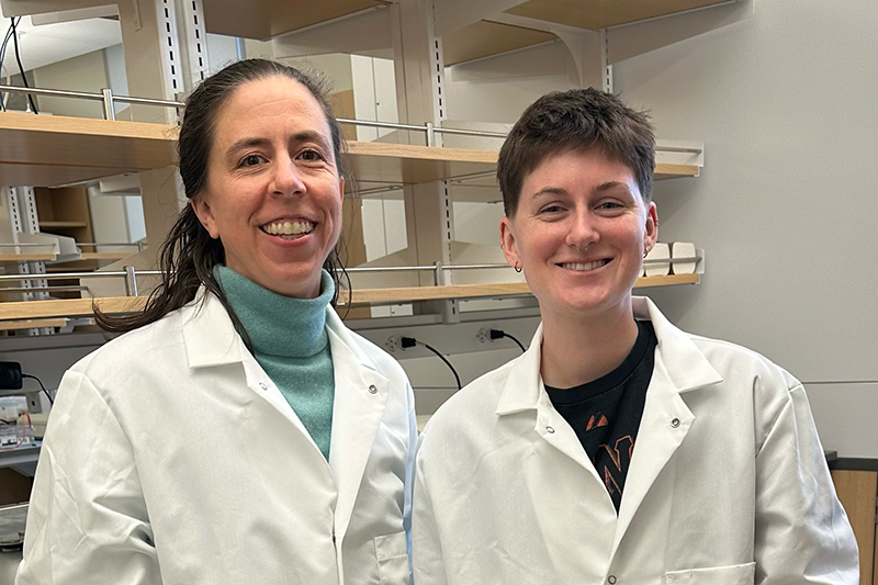 Two smiling college researchers dressed in white lab coats standing inside a research laboratory.