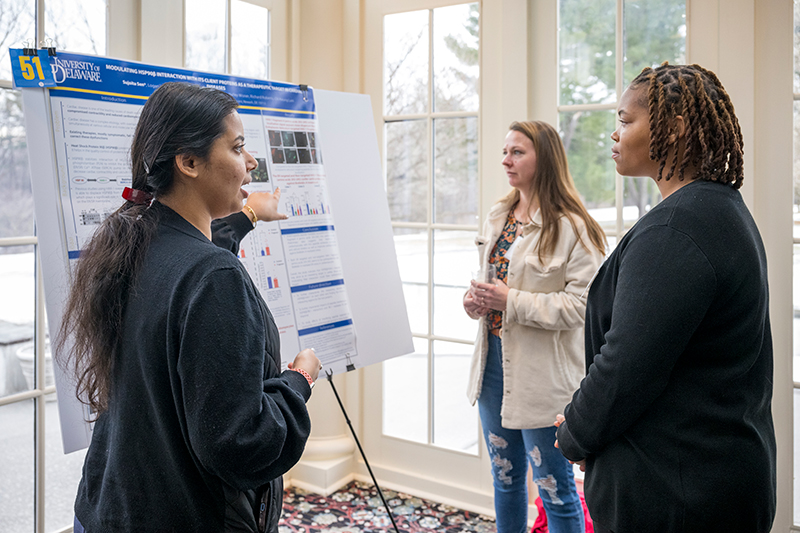 Two people listen attentively to a college student who presents her research while standing next to a poster on an easel.