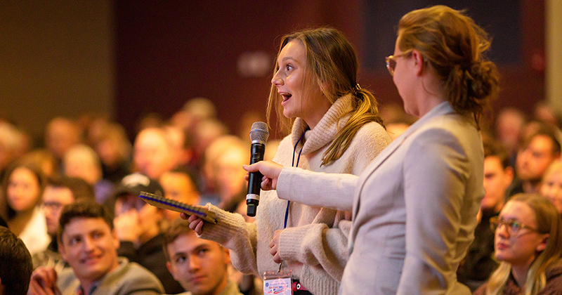 Two female University of Delaware students attending an event inside Mitchell Hall's theater. One student holds a microphone for the other so she can address a question to the guest speaker.
