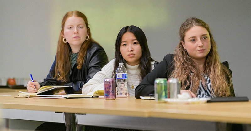 Three college students seated behind a conference table attend a meeting, listening and writing in notebooks.