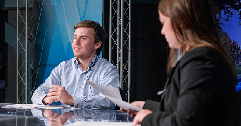 Two college students dressed in business attire sit at a news desk inside a broadcasting studio. One student faces the camera and the other student checks her notes.