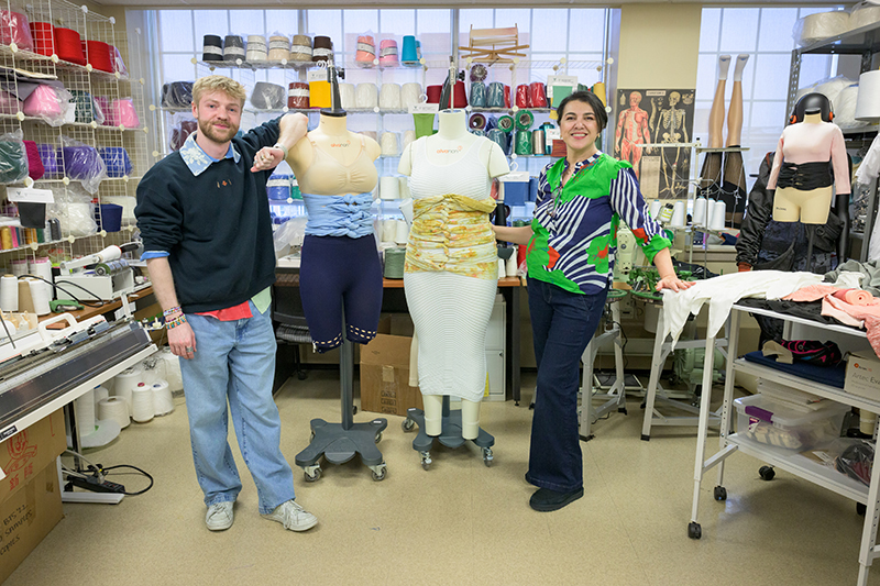 Two people in a workroom with sewing equipment and large spools of thread stand next to mannequins wearing belly band garments.