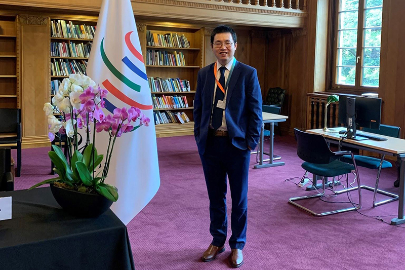 Man in business suit inside a room with a bookcase, meeting table and flag.