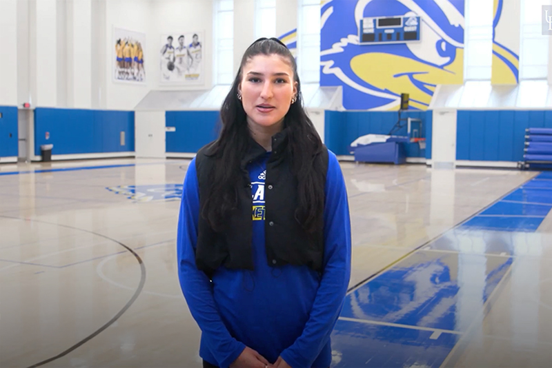 A college student in a blue UD sweatshirt standing inside a gymnasium