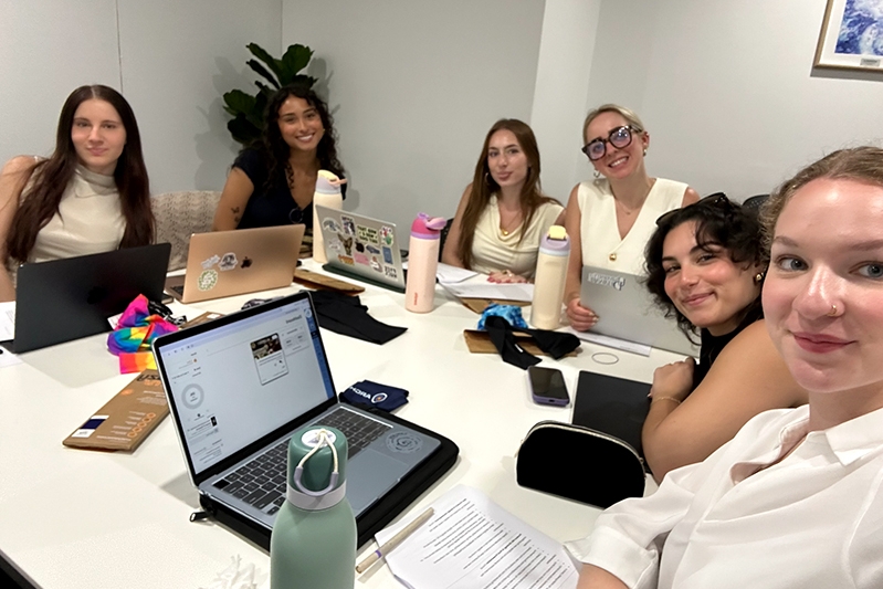 Six college students siting around a table in a conference room with laptops and water bottles. 