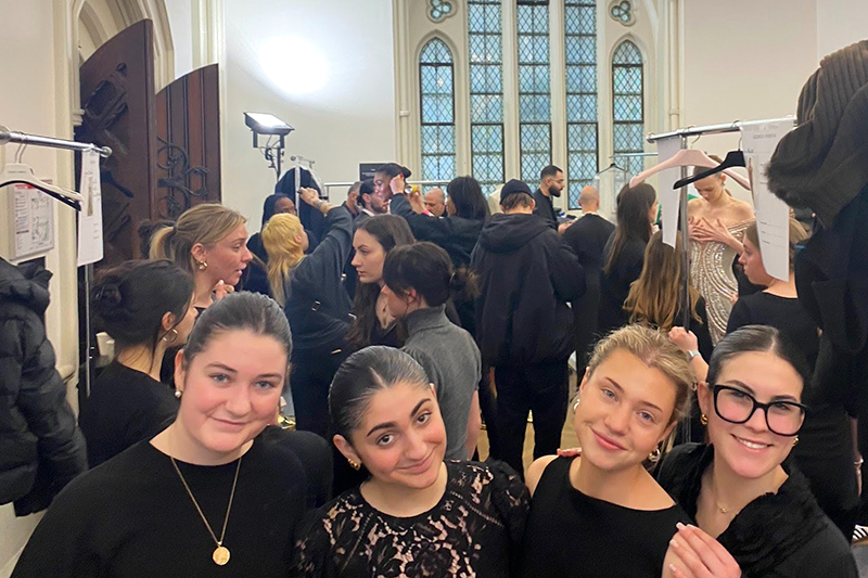 Three college students in black outfits in the foreground of a crowded room of people and two clothes racks..
