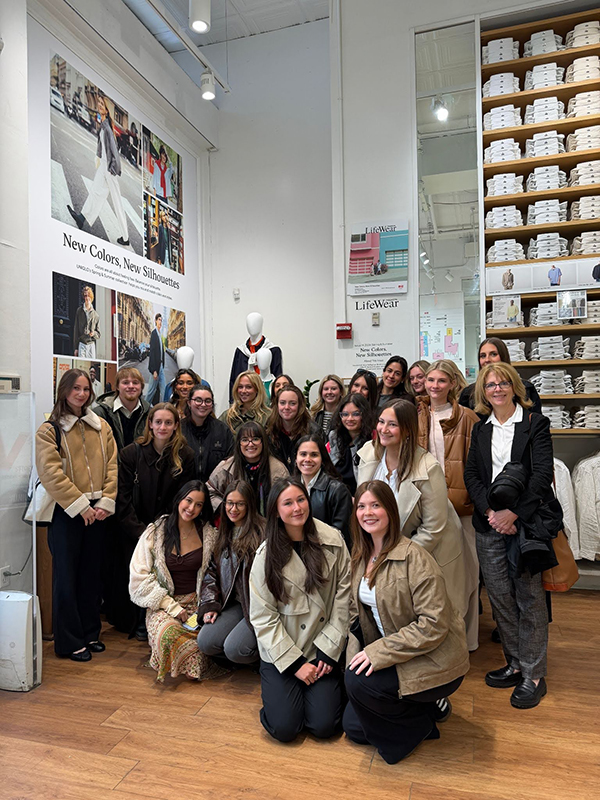 A large group of college students and faculty inside a room with posters, a mannequin and a tall shelf of folded clothing.