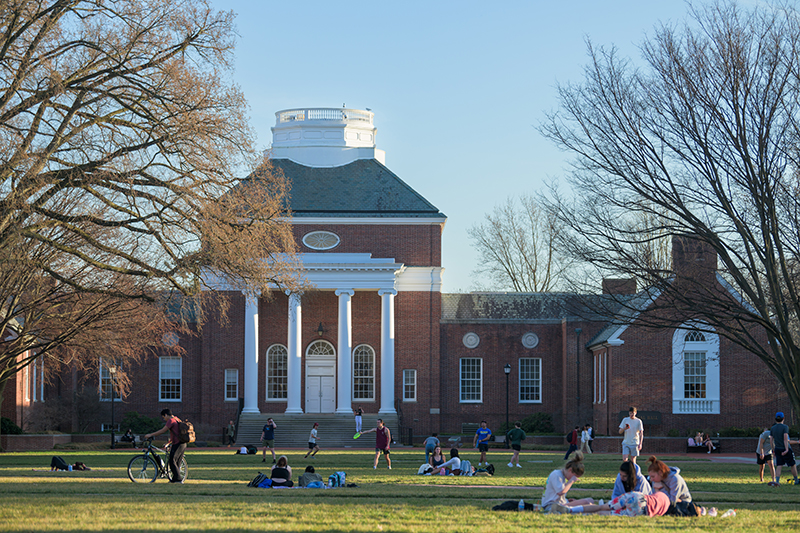 College students gather for picnics and games on a large green lawn in front of a historic brick academic building.