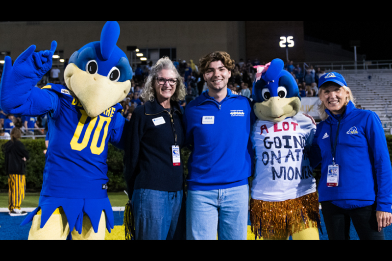 Two UD mascots and three smiling people at a night time football game inside a college stadium.