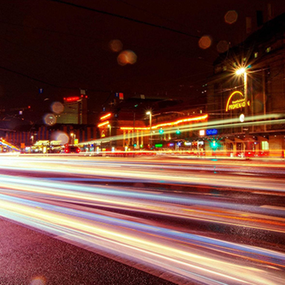 A city street at night in Leipzig, Germany, lit by colorful neon signs, traffic lights, and passing traffic.
