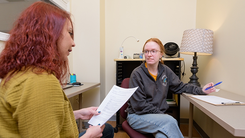 Two graduate students sit inside a small office and review documents during a meeting.