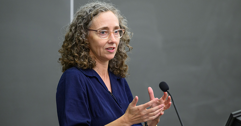 A college professor stands at the front of a lecture hall in front of a microphone and delivers a speech.