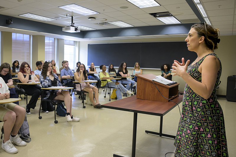 A professor stands in the front of the classroom and addresses her students.