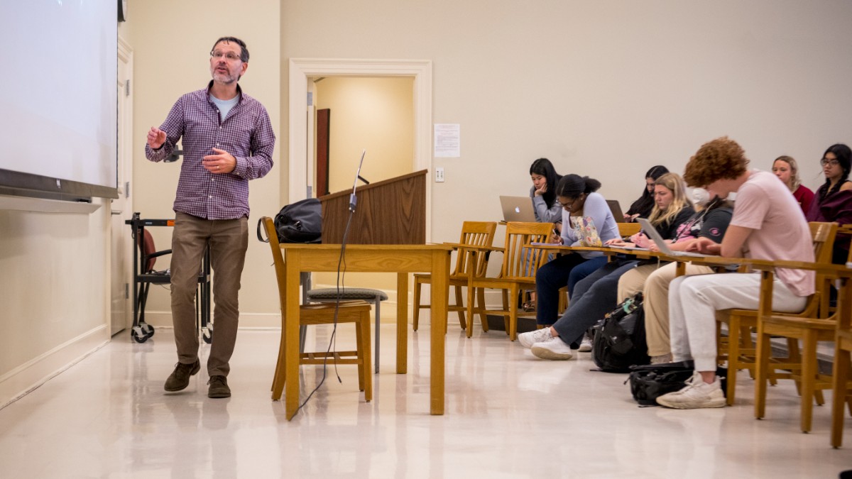 Seth Shabo, associate professor of philosophy, delivers a lecture and presents notes on a screen in the front of the classroom while students sit at their desks and take notes.